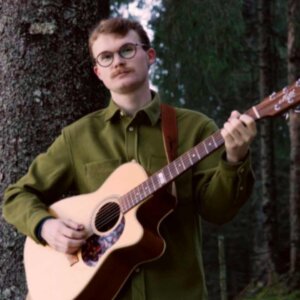 Ung mann med briller spiller gitar i skogen, kledd i grønn skjorte mot en bakgrunn av trær.