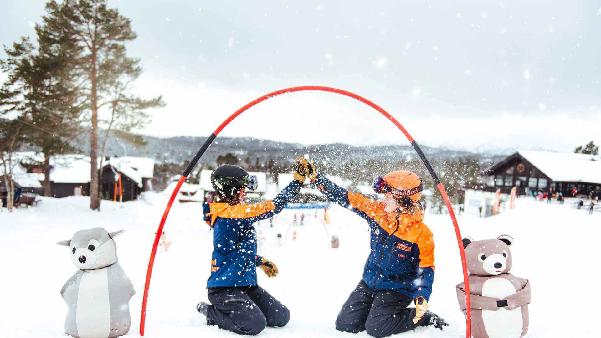 To personer i skiklær gir high-five i snøen, omgitt av snøkledde hytter og vinterpyntede figurer.