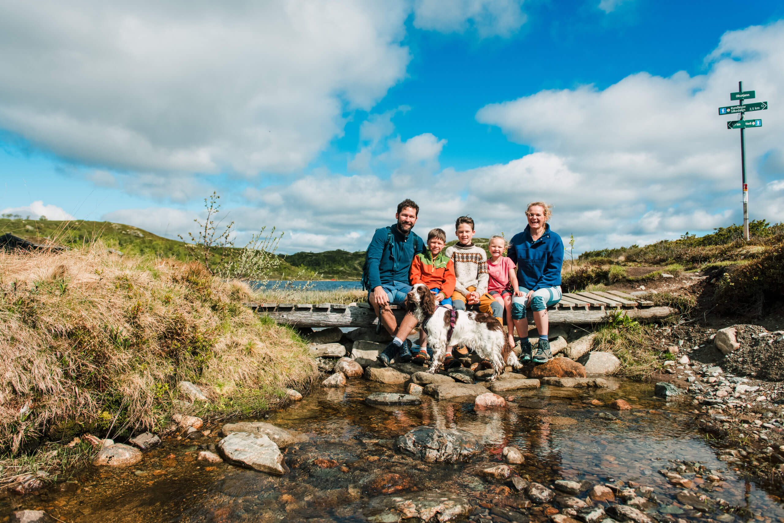 Familie med barn og hund sitter på en bro ved en elv, omgitt av natur og blå himmel. En inspirerende utendørsopplevelse.
