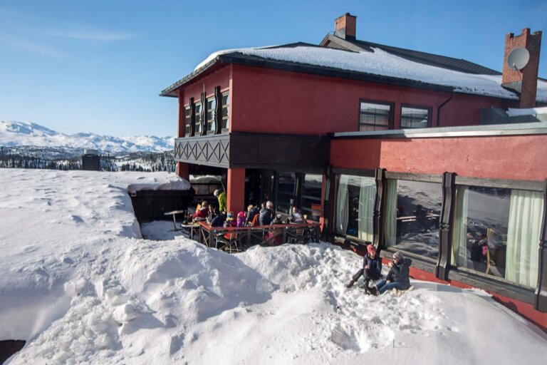 Koselig restaurant i fjellene, omringet av snø, med gjester som nyter solen og utsikten. Perfekt for après-ski.