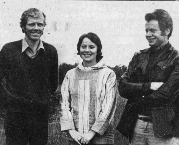 Tre personer smiler og står sammen ute på en eng. Vintage svart-hvitt bilde med en nostalgisk stemning.