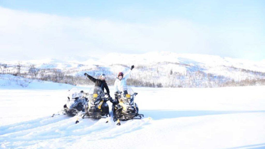 To personer poserer glade ved snøscootere i et snødekt landskap med fjell i bakgrunnen.