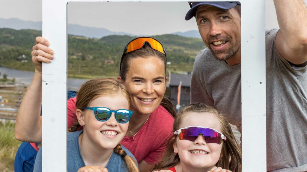 Familieportrett med to voksne og to barn med solbriller, smilende foran en landskapsutsikt i Norge.