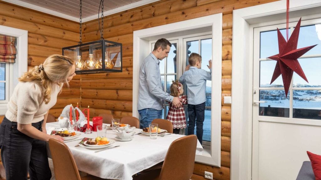 Familie nyter tid sammen i en hytte med utsikt over snødekte landskap, bord dekket for måltid, julepyntet rom.