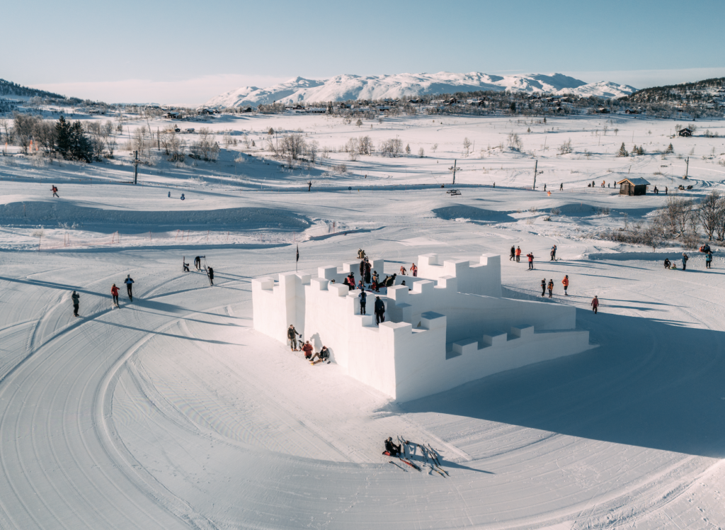 Snøslottslek i vinterlandskap med fjell i bakgrunnen og blå himmel. Barnefamilier nyter vinteraktiviteter.