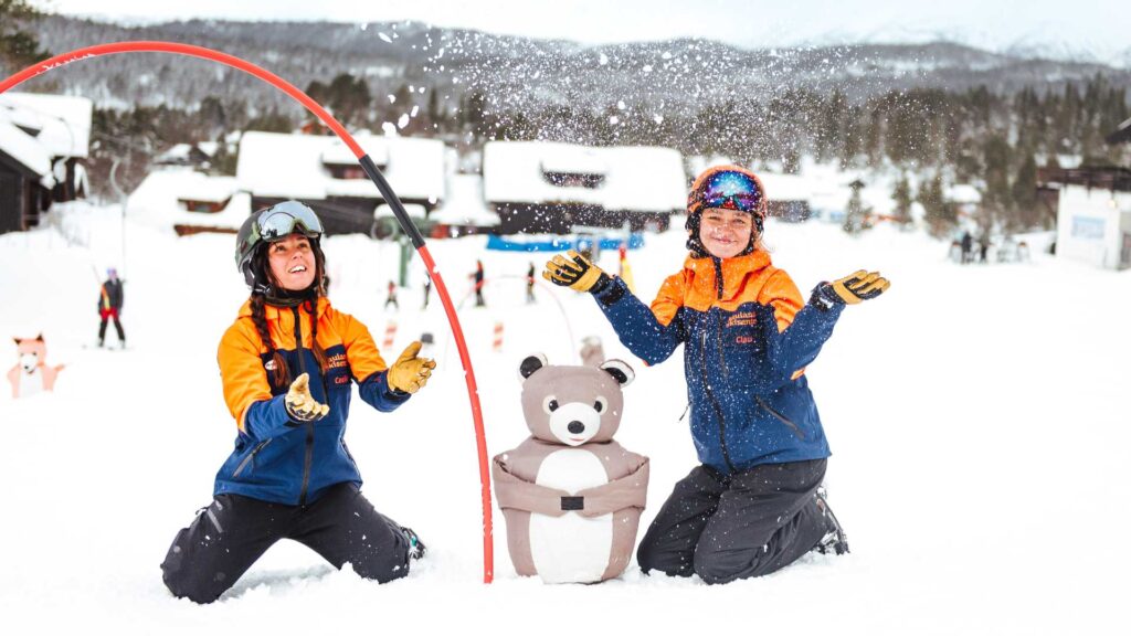 To skigledeinstruktører på snødekt bakke med bamsefigur, skaper vintermoro for barn i alpinanlegg.