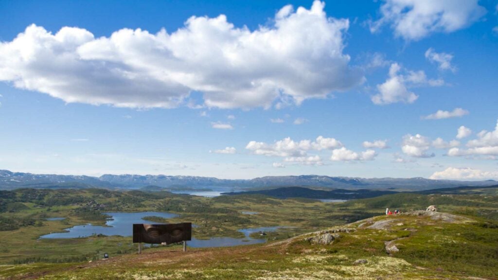 Vidstrakt landskap i Norge med fjell, innsjøer og en monumental skulptur under blå himmel og skyer.