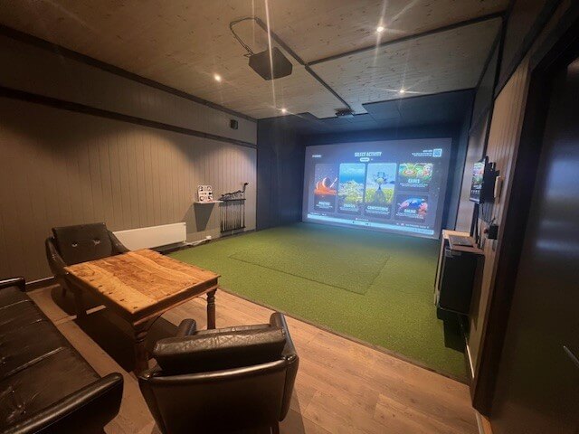 Golf-simulator i et koselig rom med stoler, bord og stor skjerm for innendørs golfsimulering.